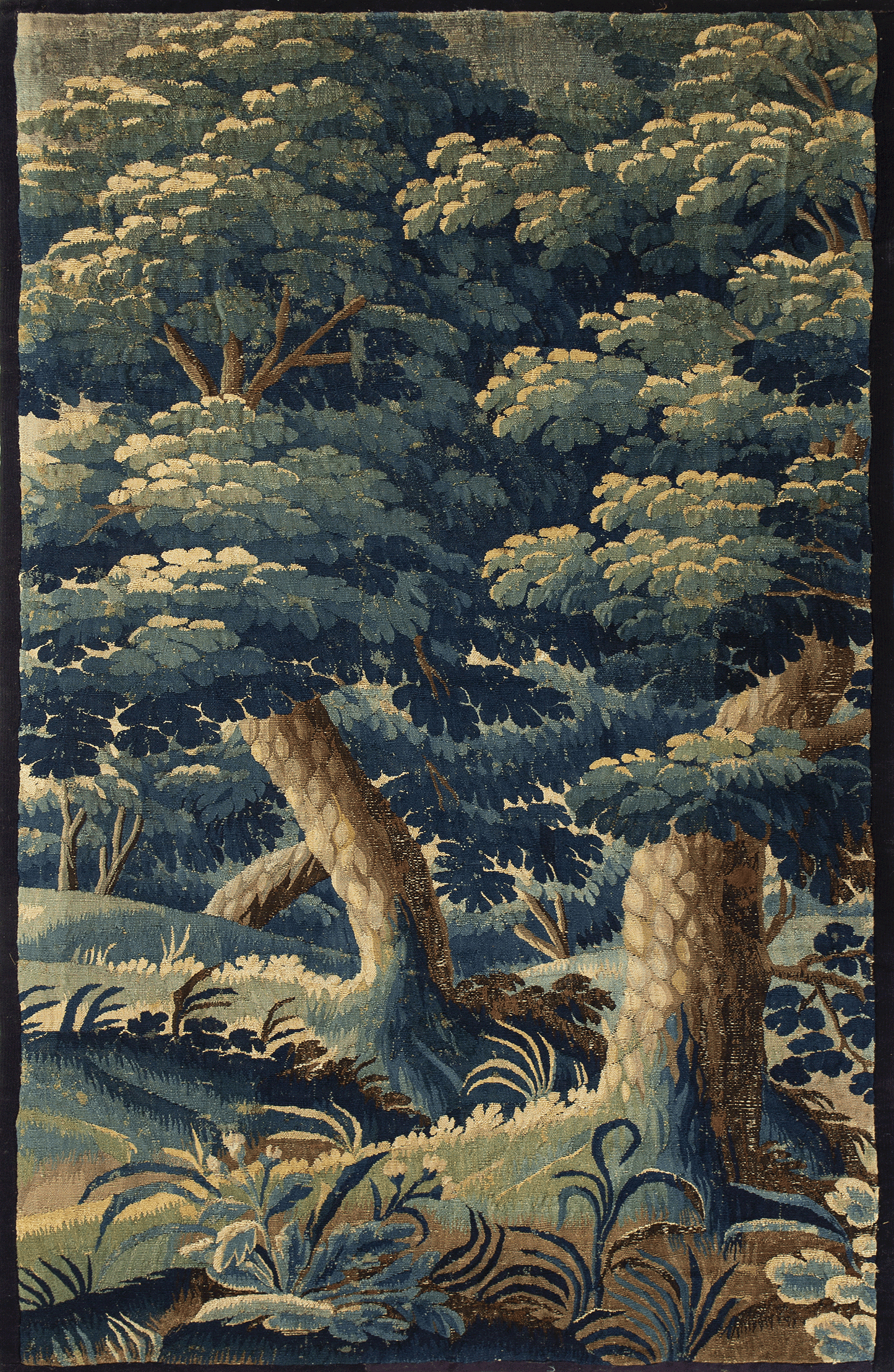 This verdure tapestry depicts a lush and detailed forest scene, rich with varying shades of green. The foreground is dominated by an intricate display of flora, with leaves and fronds meticulously woven to create depth and texture. Tall trees with thick canopies rise majestically, their trunks and branches exhibiting a naturalistic style and strong vertical lines that draw the eye upwards. 